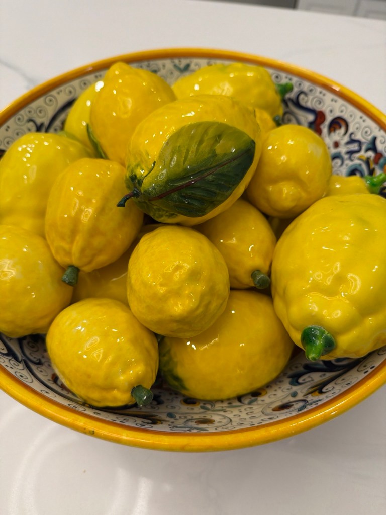 Glossy yellow ceramic lemons, some with green leaves, piled in a patterned ceramic bowl with a yellow rim on a white tabletop.