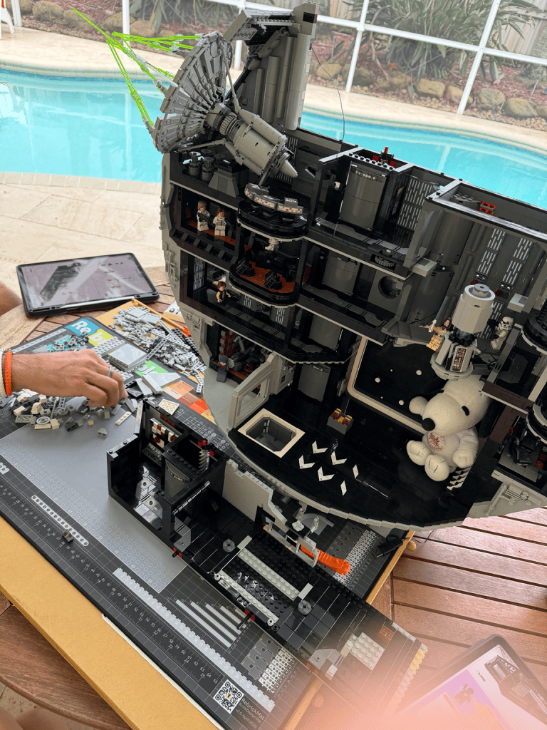 LEGO build in progress on an outdoor table beside the pool, with loose bricks, digital instructions and Snoopy tucked inside the partially completed build.