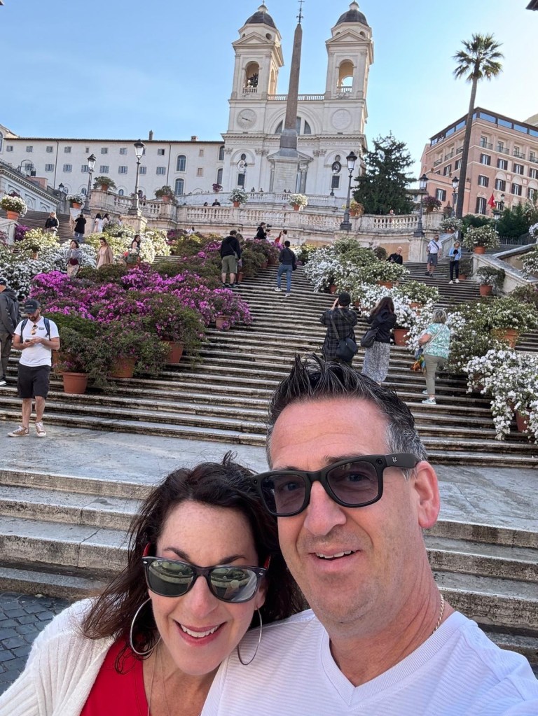 Wendi and Dan smiling at the Spanish Steps in Rome, with pink and white flowers and the twin-towered church above them.