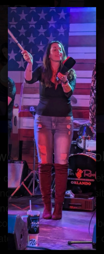 Wendi smiling and playing cowbell onstage during a live On the Rocks performance.