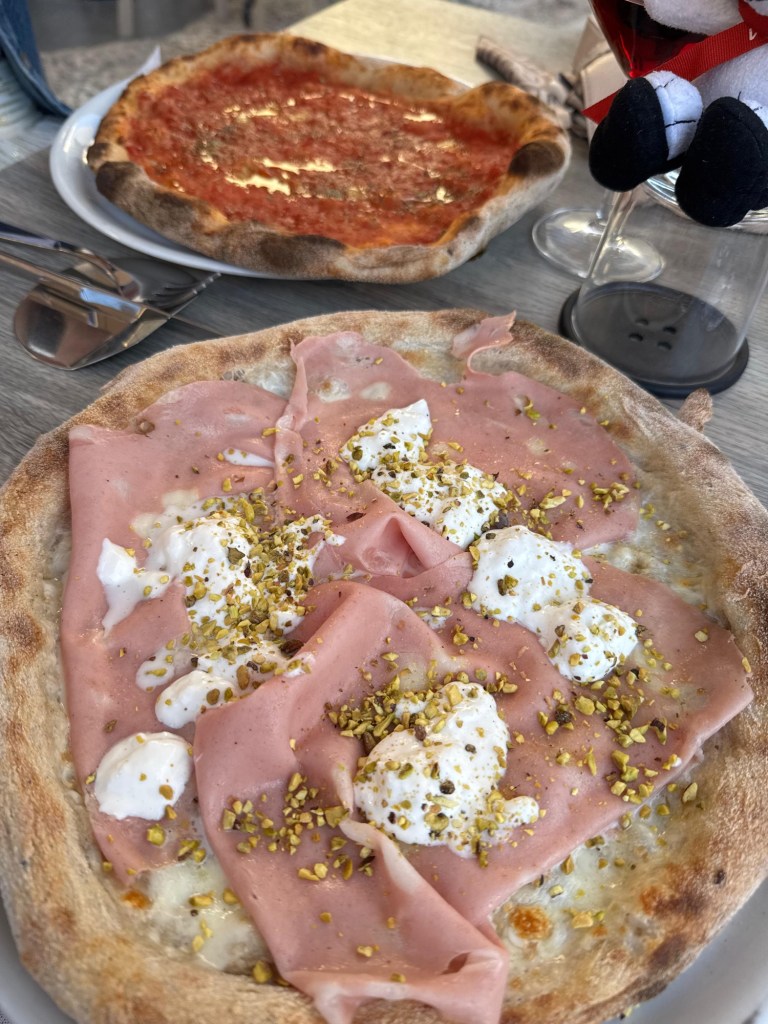 Two pizzas on a restaurant table in Monopoli: one topped with sliced meat, white cheese, and chopped nuts, and the other with tomato sauce. Snoopy peeks into the corner.