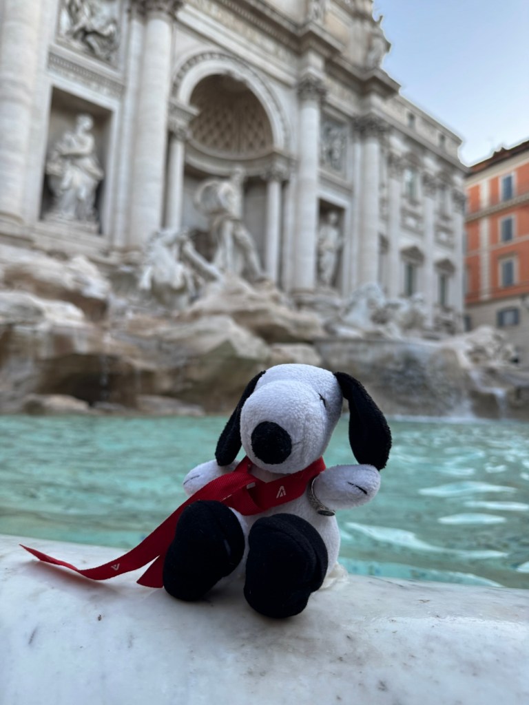 Snoopy plush wearing a red ribbon, perched beside the turquoise water of Rome’s Trevi Fountain, with marble sculptures behind him.