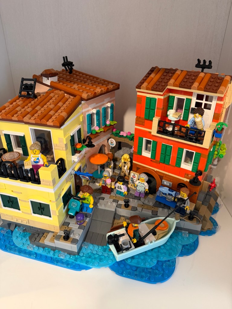 Colorful LEGO Italian village scene with waterfront buildings, balconies, café tables, flowers, minifigures and a small boat.