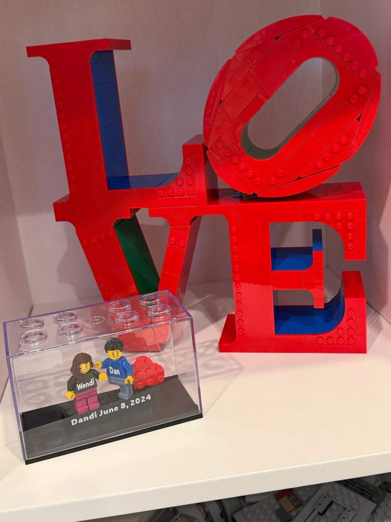 LEGO minifigures of Wendi and Dan displayed together in front of a large red LOVE LEGO sculpture, commemorating their June 8, 2024 wedding.