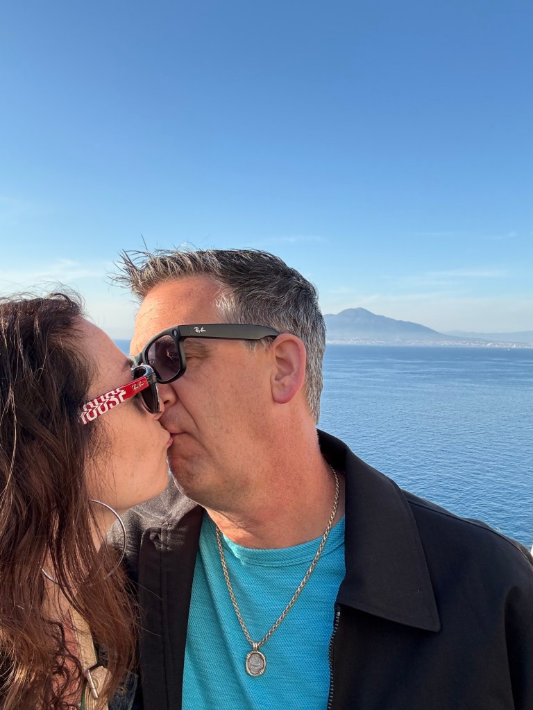 Wendi and Dan sharing a kiss with blue sea and distant mountains behind them.