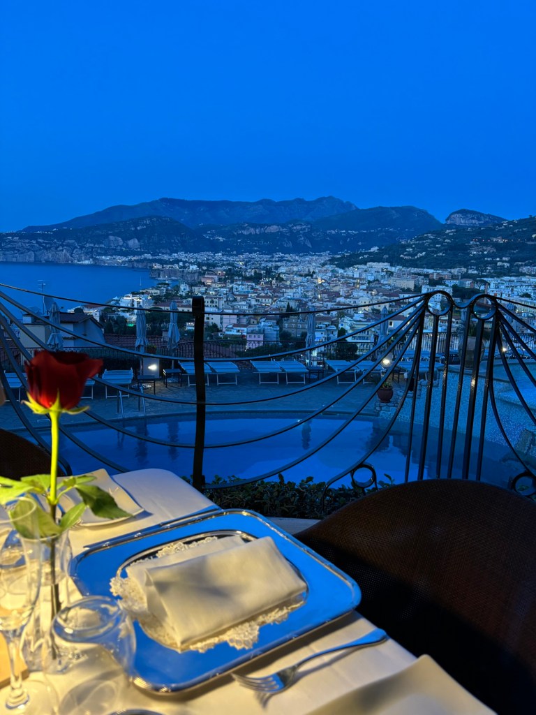 Sorrento at twilight, with the sea, coastal buildings and mountains beyond a terrace set for dinner.