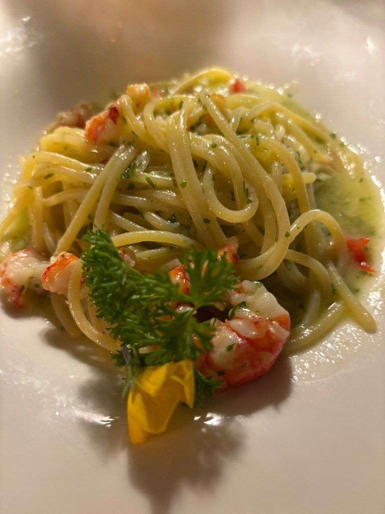Long pasta with pieces of seafood, green herbs, and a yellow flower garnish in a white bowl.