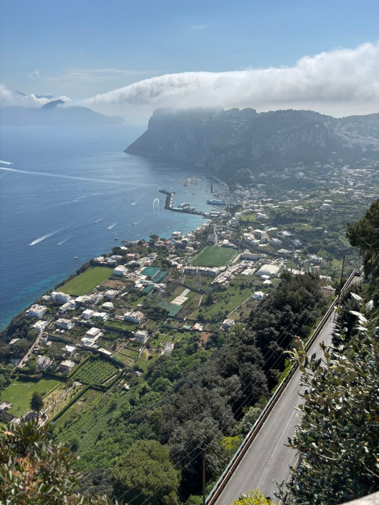 View over Capri’s coastline and harbor, with blue sea, white buildings, green gardens, and low clouds spilling over distant cliffs.