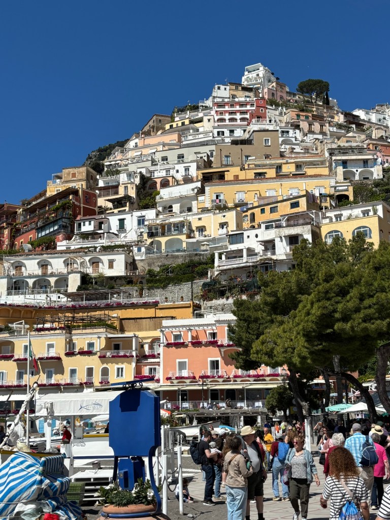 Colorful buildings climbing the steep hillside in Positano, with visitors walking below under a bright blue sky.