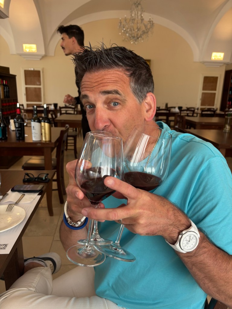Dan smiling and holding three glasses of red wine at a tasting, wearing a turquoise shirt, with wine bottles and tables behind him.