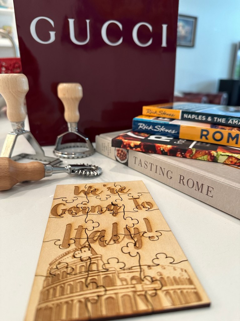 Wooden birthday puzzle reading “We’re Going to Italy!” with an illustration of the Colosseum, beside Italian travel books and pasta-making tools.