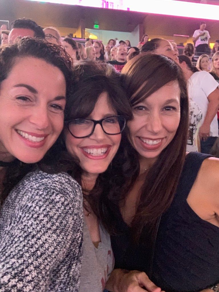 Wendi and two friends smiling together in the crowd at a concert.