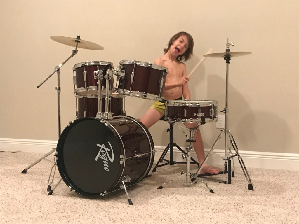 AJ as a young child enthusiastically playing a full drum set at home.